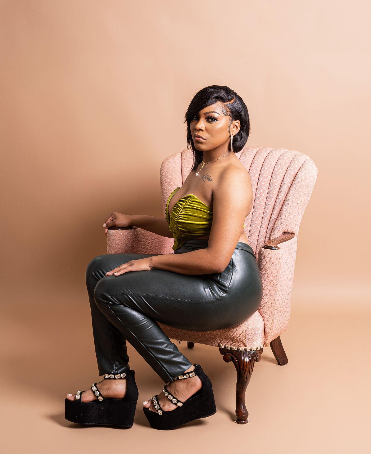 Person wearing a green top and black leather pants, sitting on a vintage pink chair.